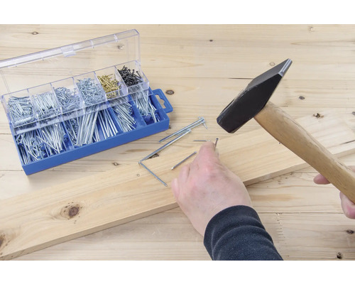 Ciocanul bate un cui într-o scândură de lemn lângă cutia de sortimente cu cuie.