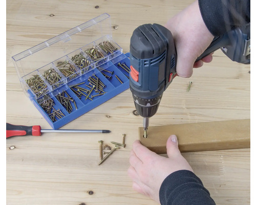 Mașină de înșurubat cu acumulator înșurubând un șurub într-o bucată de lemn, sortiment de șuruburi și șurubelniță în fundal