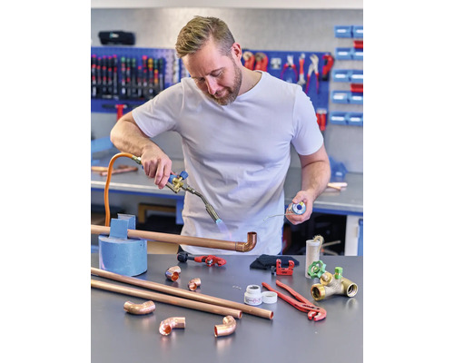 Un instalator sudează țevi de cupru cu un arzător, în timp ce diverse scule și piese de țevi sunt așezate pe bancul de lucru.