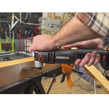 O persoană folosește un instrument multifuncțional marca Worx pentru a prelucra o bucată de lemn. Instrumentul este negru și portocaliu.