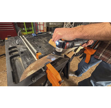 O persoană folosește un instrument multifuncțional pentru șlefuirea unei piese de lucru, care este fixată cu cleme pe un banc de lucru.