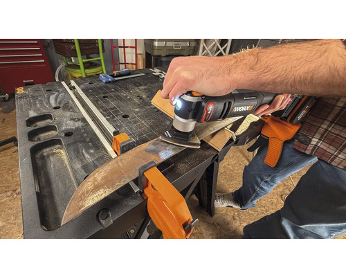 O persoană folosește un instrument multifuncțional pentru șlefuirea unei piese de lucru, care este fixată cu cleme pe un banc de lucru.