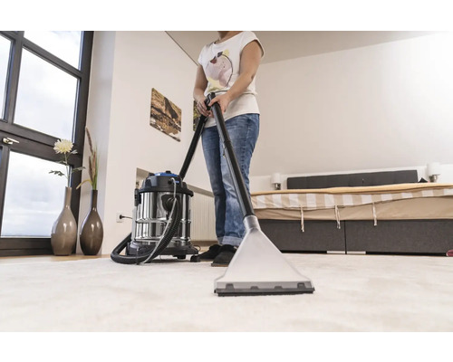 Femeie curăță covorul cu un aspirator cu injecție-extracție într-un dormitor luminos.