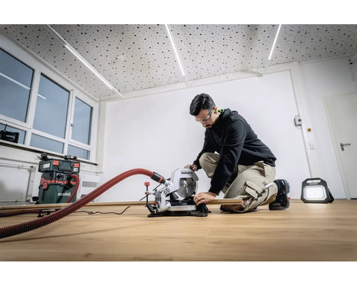Un bărbat îngenuncheat manevrează un fierăstrău circular Metabo conectat la un aspirator. În fundal se văd ferestre și o lampă de lucru.