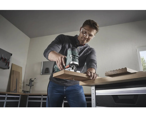 Un bărbat cu ochelari de protecție folosește un ferăstrău vertical pentru a tăia o bucată de lemn. Atelier în fundal.