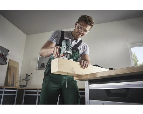 Un bărbat cu ochelari de protecție taie o scândură de lemn cu un ferăstrău vertical pe un banc de lucru.