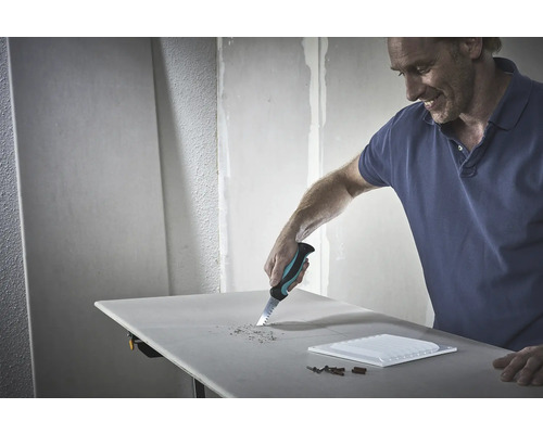 Un bărbat taie o gaură într-o placă de gips-carton cu un ferăstrău manual. Unelte și o faianță sunt așezate pe masă.