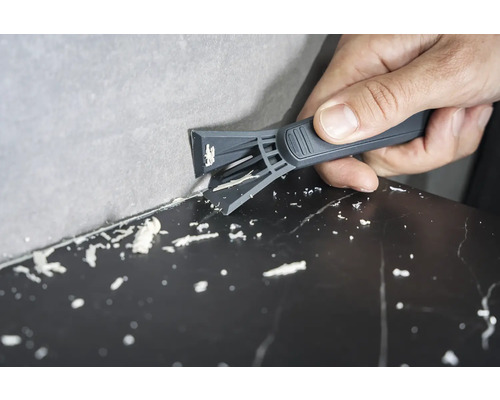 O persoană îndepărtează rosturile de silicon dintre perete și suprafață cu ajutorul unei răzuitoare pentru rosturi.