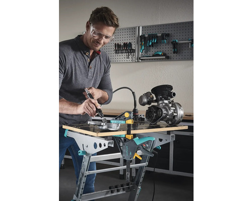 Bărbat cu ochelari de protecție prelucrează o piesă metalică pe un banc de lucru Wolfcraft cu o unealtă multifuncțională.