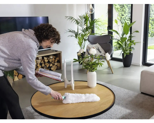 Femeie ștergând praful de pe o masă de lemn într-un living cu un pămătuf