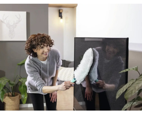 Femeie curăță un televizor cu ecran plat cu un pămătuf într-un living luminos.