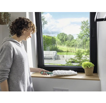 Femeie șterge pervazul ferestrei cu un pămătuf de praf. Fereastră cu vedere la verdeață.