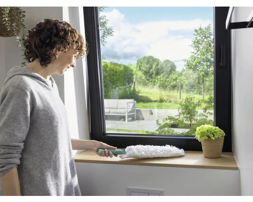 Femeie șterge pervazul ferestrei cu un pămătuf de praf. Fereastră cu vedere la verdeață.