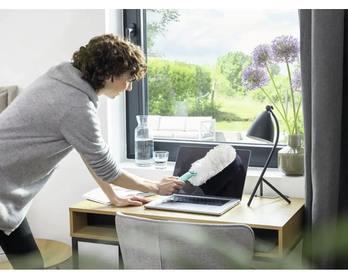 Femeie curăță laptopul cu un pămătuf pe birou.