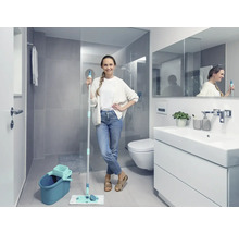 Baie luminoasă cu femeie ținând un mop și o găleată, lângă un dulap de lavoar, o toaletă și un duș.