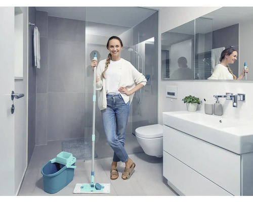 Baie luminoasă cu femeie ținând un mop și o găleată, lângă un dulap de lavoar, o toaletă și un duș.