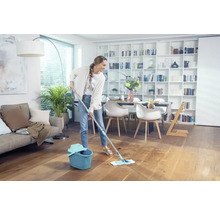 Femeie șterge podeaua de lemn cu mop și găleată într-un living luminos.