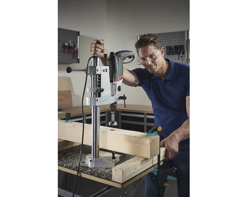 Un bărbat găurește o bucată de lemn cu o mașină de găurit de masă. Poartă ochelari de protecție.