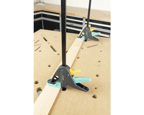 Două cleme de fixare cu o singură mână fixează o scândură de lemn pe un banc de lucru.