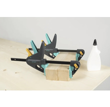 Două cleme de fixare cu o mână Wolfcraft fixează bucăți de lemn pe un banc de lucru, alături se află o sticlă de adeziv.