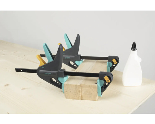 Două cleme de fixare cu o mână Wolfcraft fixează bucăți de lemn pe un banc de lucru, alături se află o sticlă de adeziv.