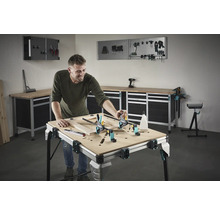Un bărbat lucrează la un cadru de lemn pe un banc de lucru într-un atelier. Unelte și echipamente de atelier sunt vizibile în fundal.
