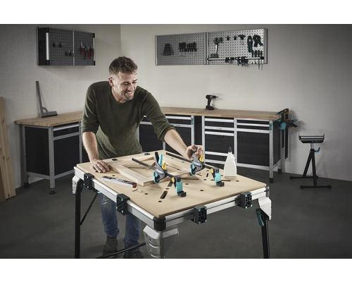 Un bărbat lucrează la un cadru de lemn pe un banc de lucru într-un atelier. Unelte și echipamente de atelier sunt vizibile în fundal.