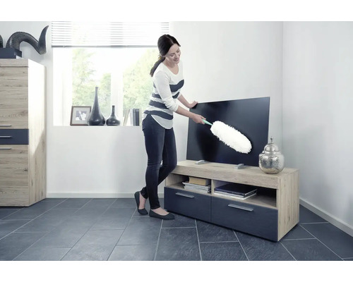 Femeie șterge praful de pe un televizor cu un pămătuf într-un living.