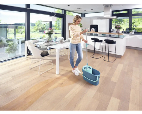 Femeie șterge podeaua de lemn cu mop și găleată într-o zonă de locuit luminoasă.