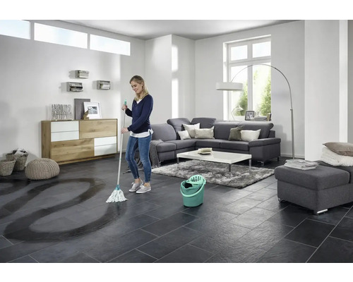 Femeie ștergând podeaua cu gresie cu mopul și găleata într-un living luminos.