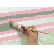 O persoană pictează o linie verde cu o pensulă rotundă pe un perete cu dungi roz și albe