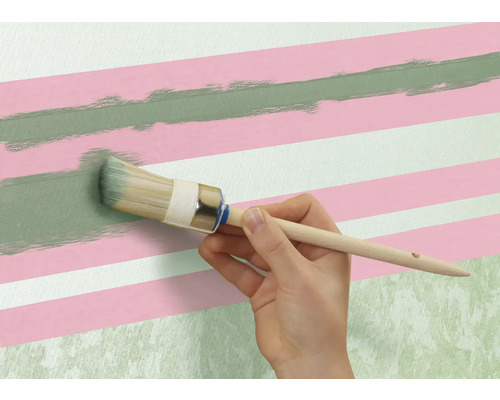 O persoană pictează o linie verde cu o pensulă rotundă pe un perete cu dungi roz și albe