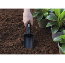 O mână ține o lopată de plantat în pământ, lângă plante în ghiveci.