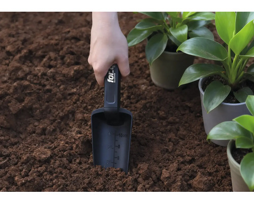 O mână ține o lopată de plantat în pământ, lângă plante în ghiveci.