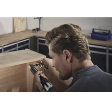 Un bărbat fixează o balama cu o șurubelniță electrică pe un dulap de lemn.