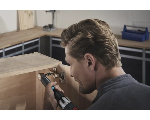 Un bărbat fixează o balama cu o șurubelniță electrică pe un dulap de lemn.