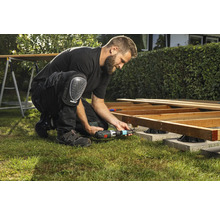 Un bărbat construiește o terasă din lemn cu o șurubelniță cu acumulator și genunchiere în grădină.