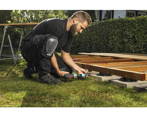 Un bărbat construiește o terasă din lemn cu o șurubelniță cu acumulator și genunchiere în grădină.