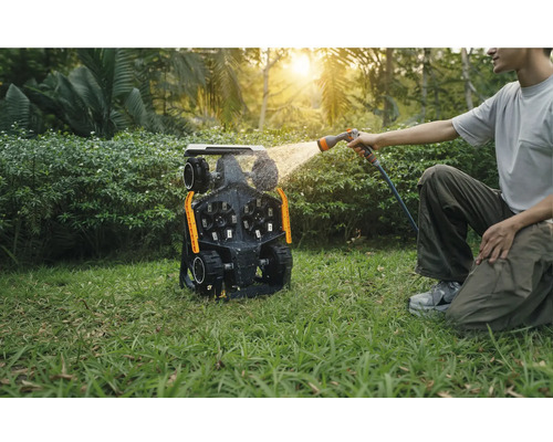 Bărbat curăță un robot de tuns iarba cu un furtun de apă în grădină.