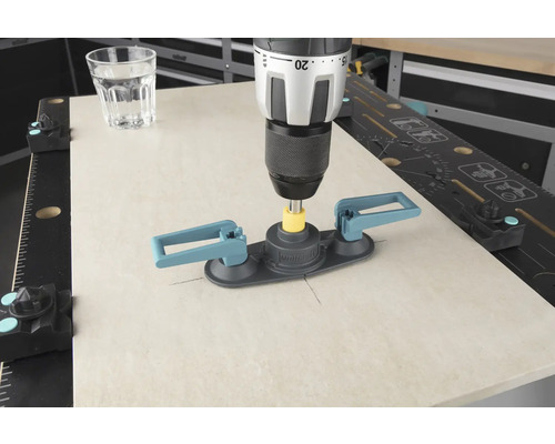 O mașină de găurit cu ajutor pentru găurirea plăcilor de faianță găurește o placă de faianță, lângă ea se află un pahar cu apă.