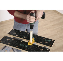 O persoană găurește cu o mașină de găurit o bucată de lemn care este fixată pe un banc de lucru cu un element de fixare galben.