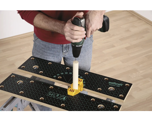 O persoană găurește cu o mașină de găurit o bucată de lemn care este fixată pe un banc de lucru cu un element de fixare galben.