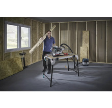 Bărbat cu grindă de lemn, ferăstrău circular și banc de lucru într-o construcție nefinisată.