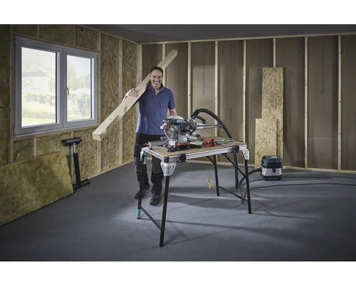 Bărbat cu grindă de lemn, ferăstrău circular și banc de lucru într-o construcție nefinisată.