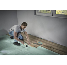 Un bărbat montează parchet laminat cu un ciocan și un bloc de lovire pe izolație fonică într-o cameră.