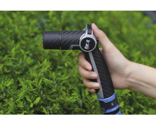 O mână ține un pistol de stropit grădina marca For Q în fața unui arbust verde