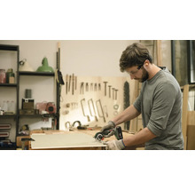 Un bărbat taie o scândură de lemn cu un fierăstrău circular portabil. Poartă ochelari de protecție și mănuși.