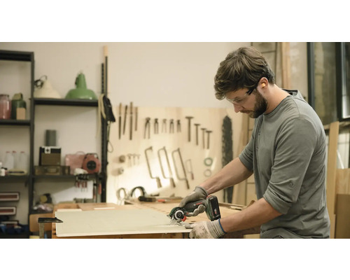 Un bărbat taie o scândură de lemn cu un fierăstrău circular portabil. Poartă ochelari de protecție și mănuși.