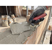 Zidar care lucrează cu o mistrie pe un zid de cărămidă, saci de ciment în fundal.