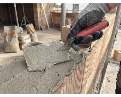 Zidar care lucrează cu o mistrie pe un zid de cărămidă, saci de ciment în fundal.